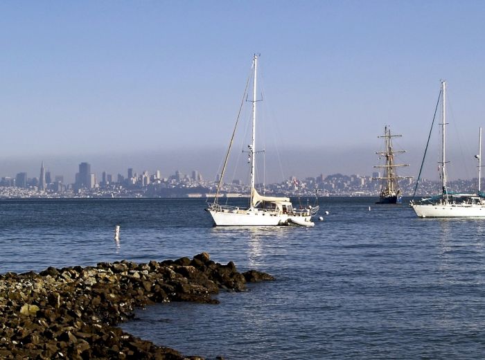 Sailboats and a tall ship are on calm waters with a rocky shoreline, and a city skyline in the background.