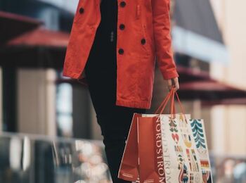 A person wearing a red coat is holding shopping bags, walking in an outdoor setting with red umbrellas in the background.