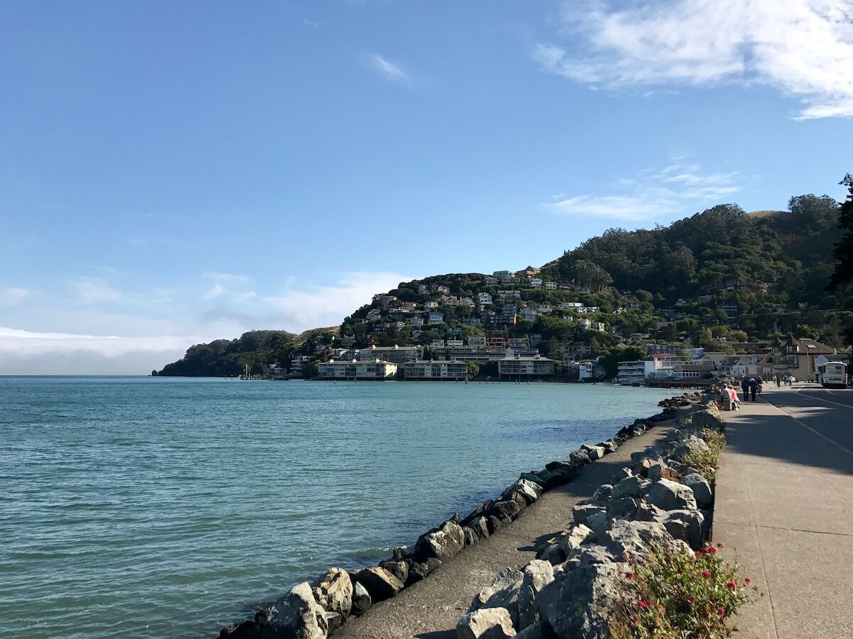 A serene coastal scene with a paved pathway alongside the water, lined with rocks, and a hillside community in the background under a clear sky.