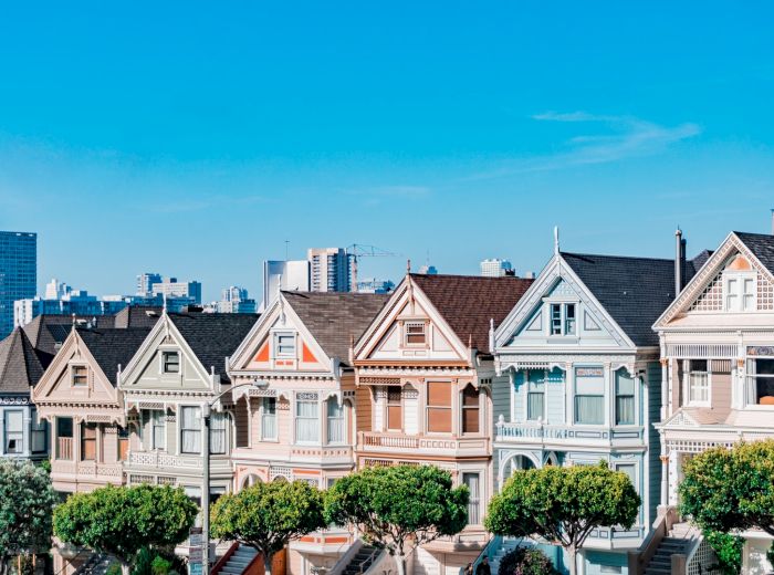The image shows a row of colorful Victorian houses with a city skyline in the background, under a clear blue sky.