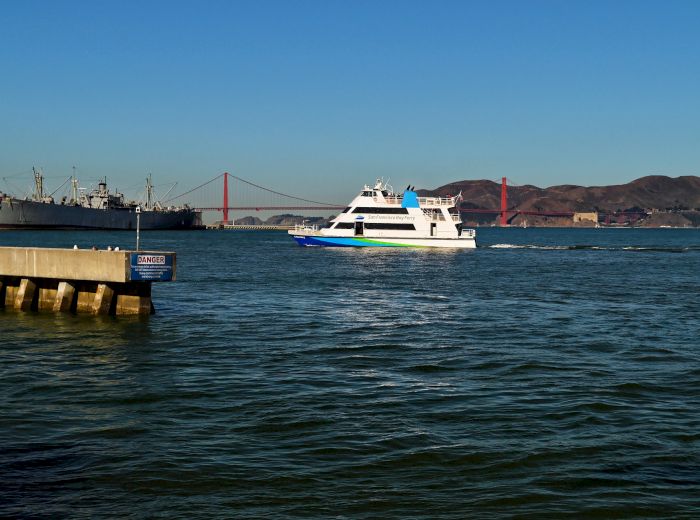 The image shows a ferry boat on the water near a dock, with a red suspension bridge and mountains in the background under a clear blue sky.