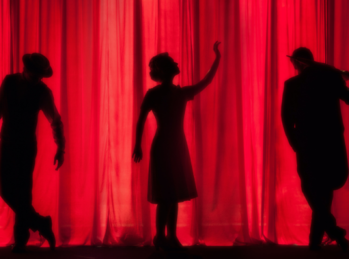 Silhouetted figures of three dancers, posed dramatically against a red curtain backdrop, create a striking theatrical scene.