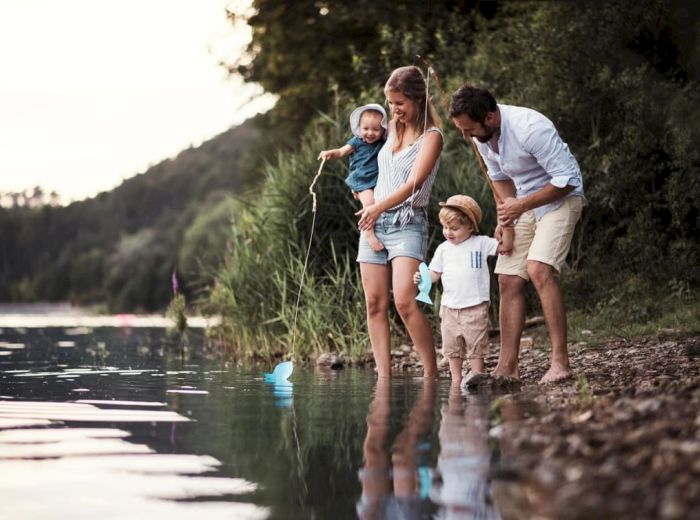 A family with two young children is standing by a lakeside, enjoying nature and the calm water, likely spending quality time together at the shore.