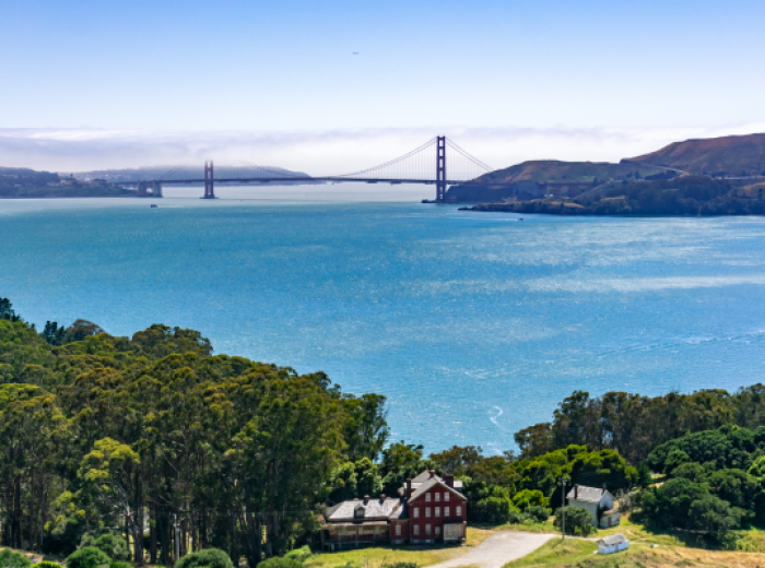 This image shows the Golden Gate Bridge spanning a bay, with clear blue water and lush greenery in the foreground, and hills in the background.