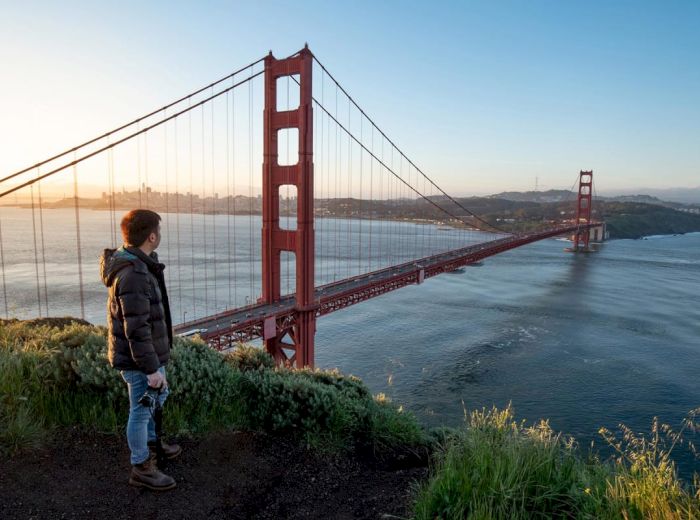 A person stands on a grassy cliff, looking at the Golden Gate Bridge stretching across the water with the city faintly visible in the background.
