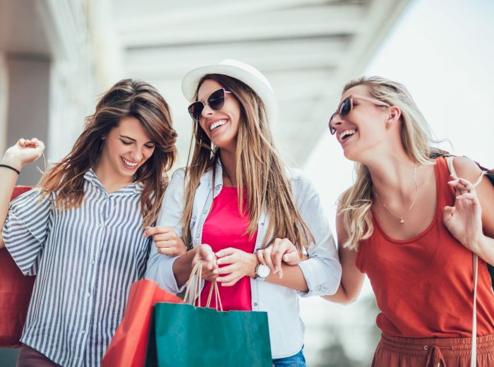 Three women are smiling and holding shopping bags while walking outside. They look happy and appear to be enjoying their time together.