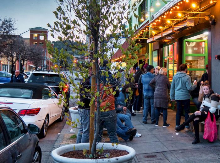 People are waiting outside a restaurant on a sidewalk with parked cars, a tree in a planter, and bright lights overhead at dusk near an intersection.