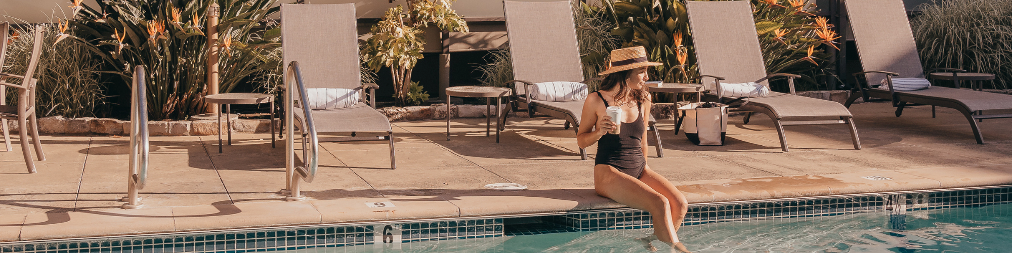 A woman in a black swimsuit and straw hat sits at the edge of a pool, with lounge chairs and plants in the background.