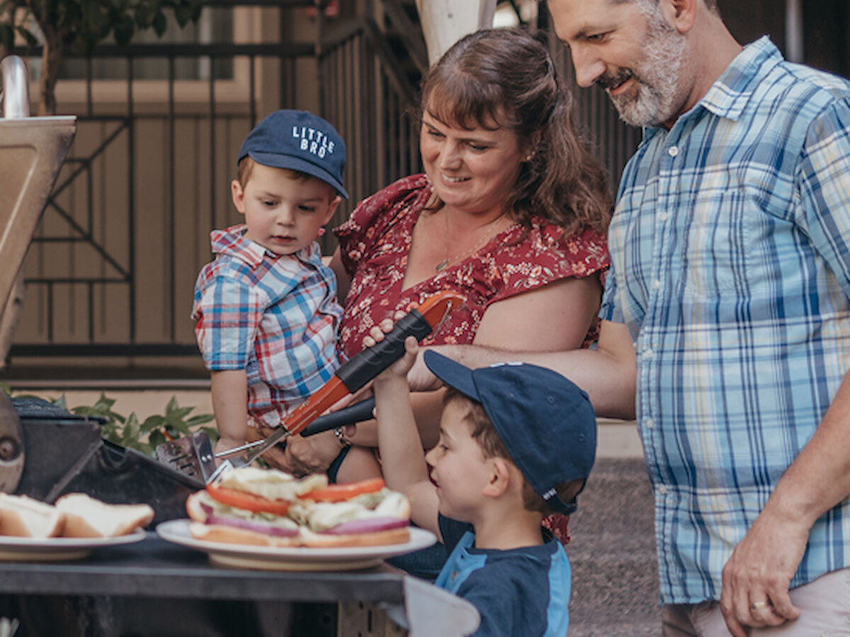 A family is gathered around a grill, with two children wearing hats and two adults smiling while preparing food outdoors.