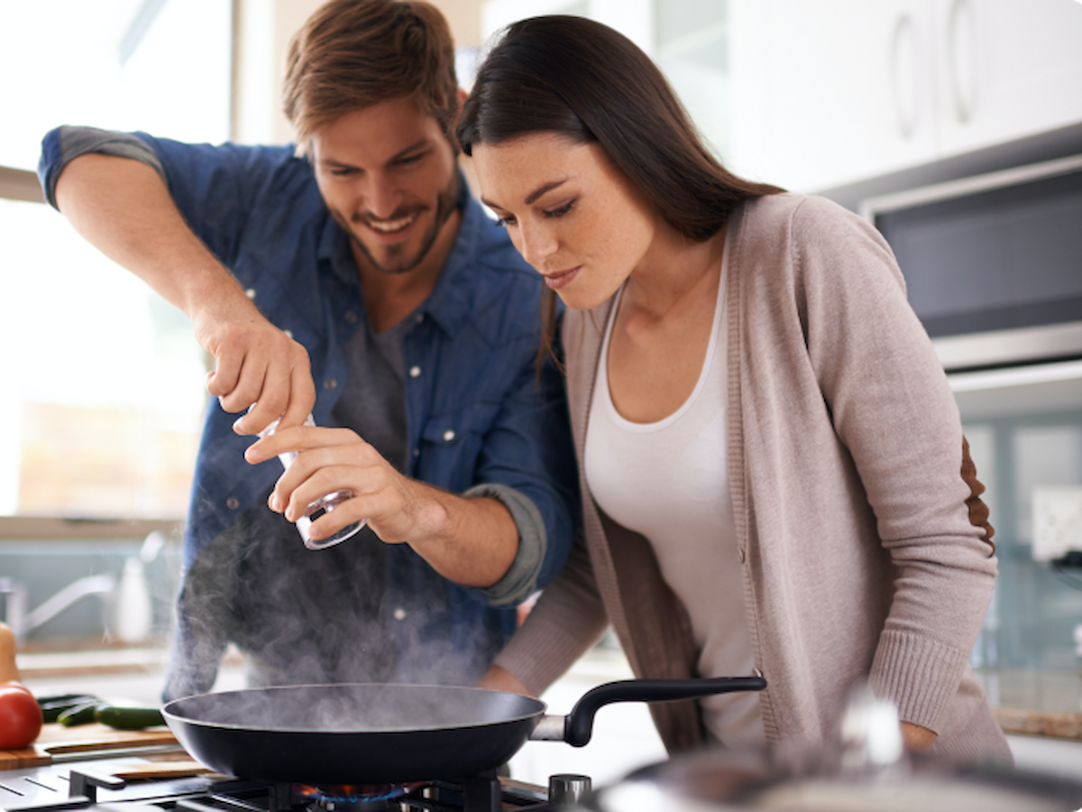 A man and woman are cooking together in a kitchen, focusing on a dish in a frying pan. The man is using a pepper grinder, and the woman is observing.