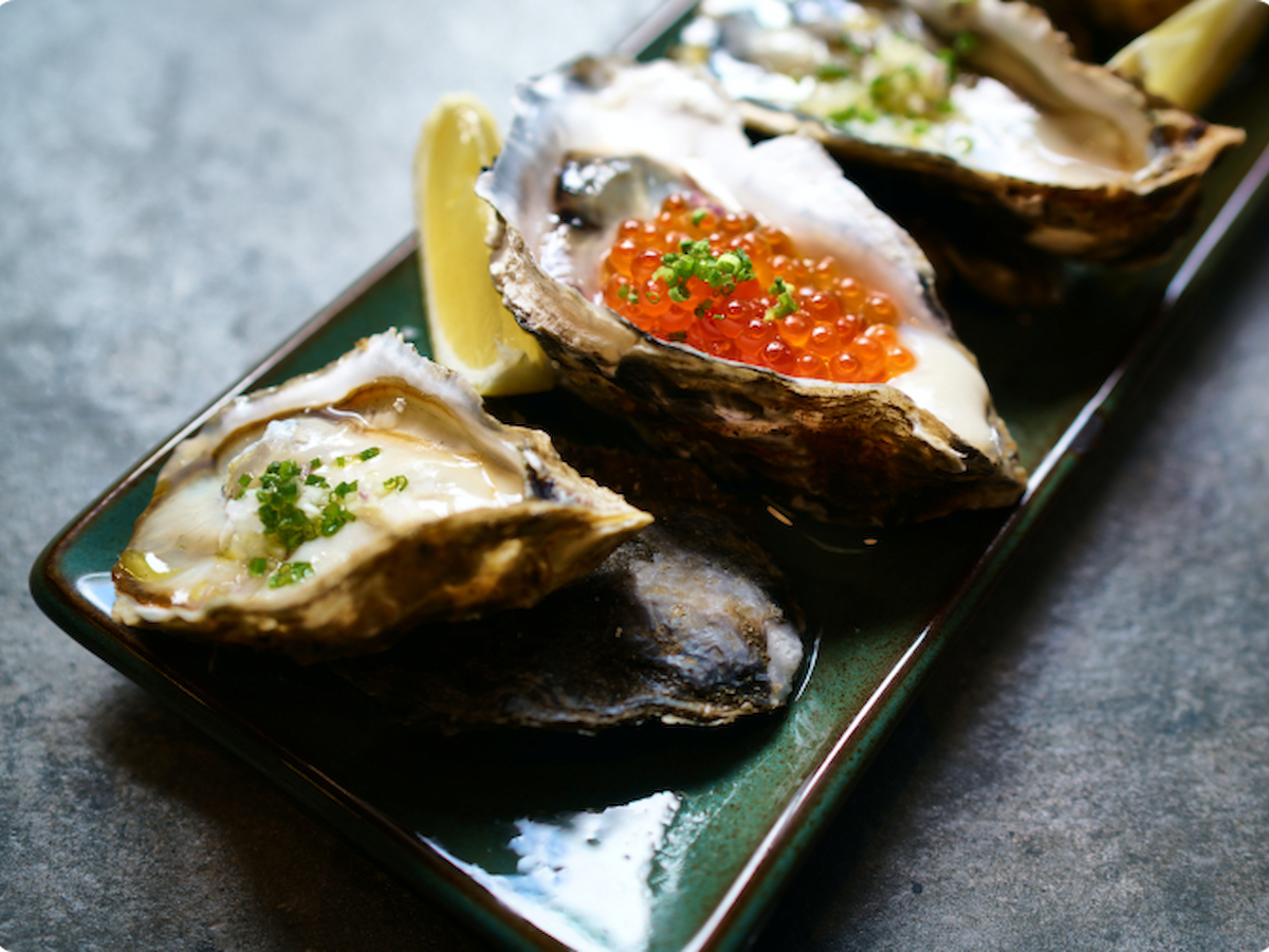 The image shows a dish of oysters on the half shell, garnished with green herbs, fish roe, and lemon wedges, served on a rectangular plate.