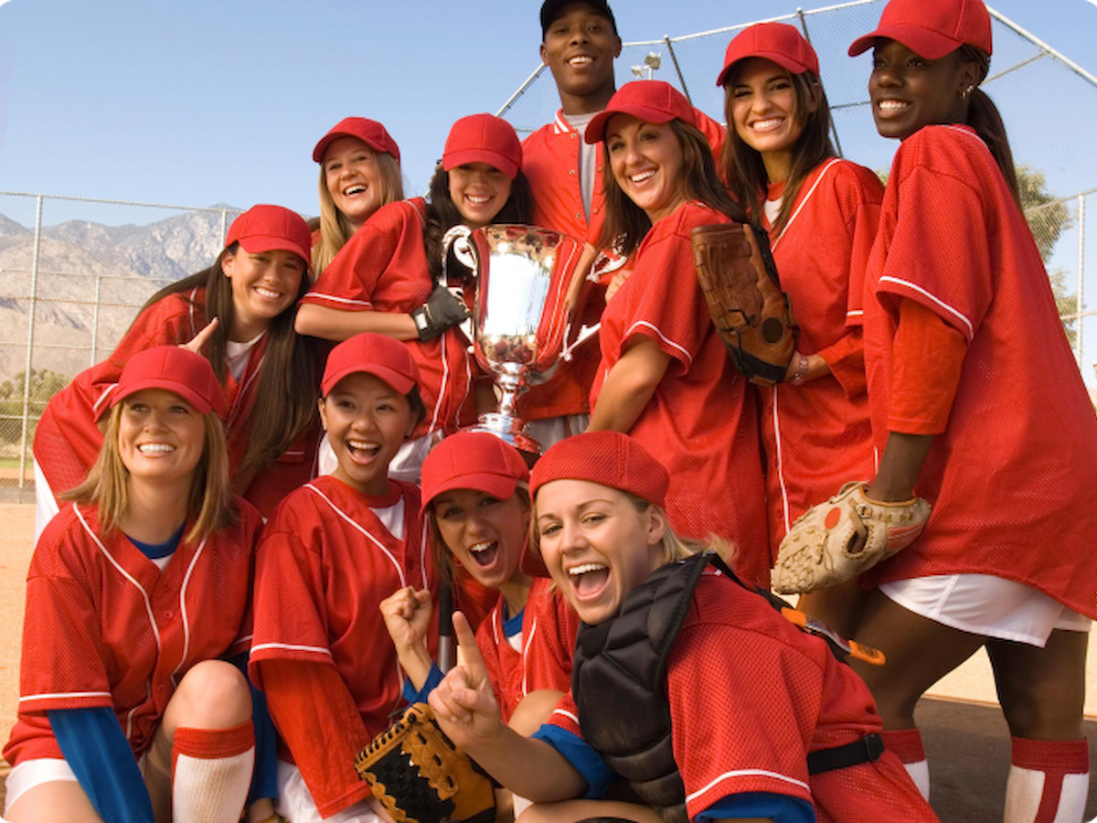 A group of people in red uniforms are celebrating on a baseball field while holding a trophy, smiling, and posing for the photo.