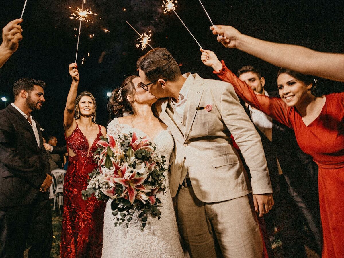 A bride and groom kiss under sparklers held by friends in celebration, featuring their wedding attire and a bouquet of flowers.