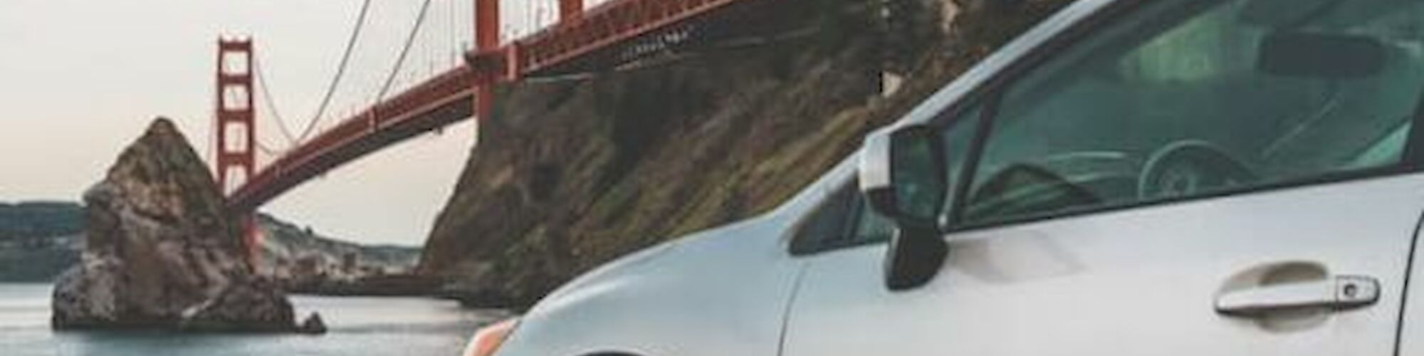 A silver car is parked near a body of water with part of the Golden Gate Bridge and a rocky island in the background.