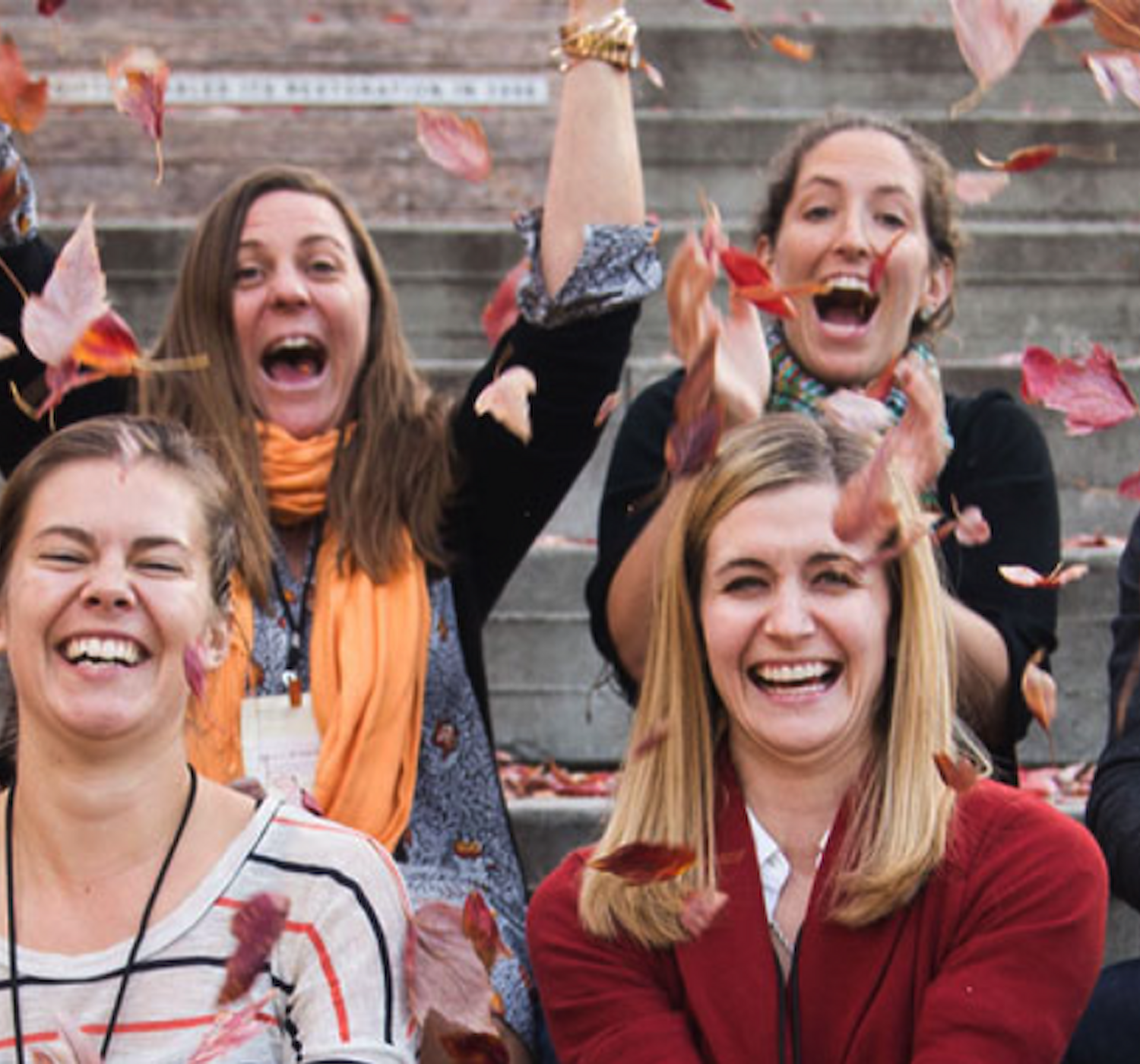 A group of people sit on stairs, smiling and laughing while tossing autumn leaves into the air, creating a joyful and playful scene.